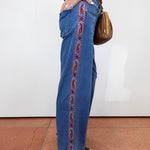 A person wearing wide-leg blue jeans with a red paisley embroidered stripe down the side, standing against a white wall on a terracotta tiled floor and carrying a brown shoulder bag.