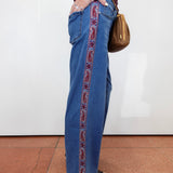 A person wearing wide-leg blue jeans with a red paisley embroidered stripe down the side, standing against a white wall on a terracotta tiled floor and carrying a brown shoulder bag.