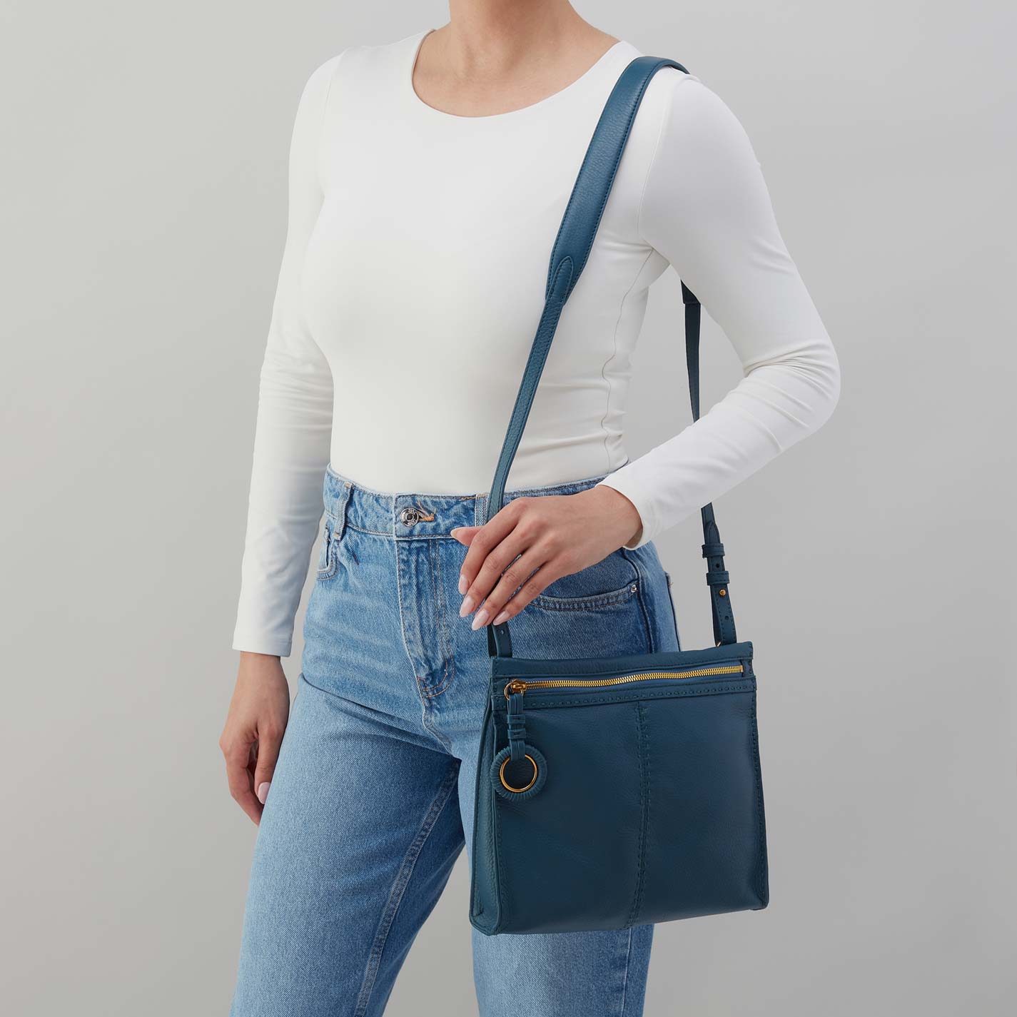 Person wearing a white long-sleeve shirt and blue jeans, holding a blue crossbody bag against a gray background