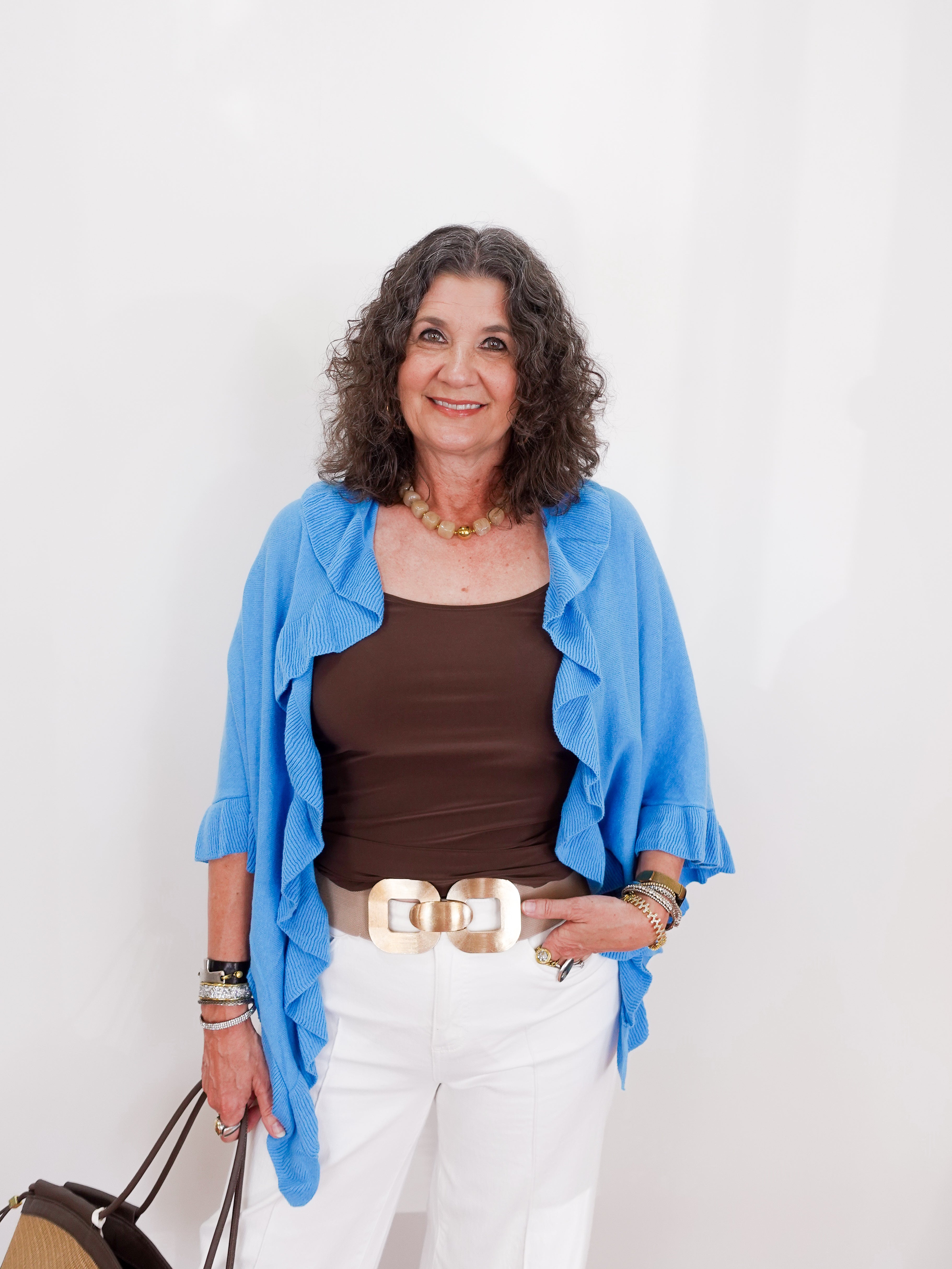 Woman wearing a blue cardigan, brown top, white pants, and carrying a handbag against a white background