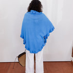 Person wearing a blue poncho and white pants against a white wall.