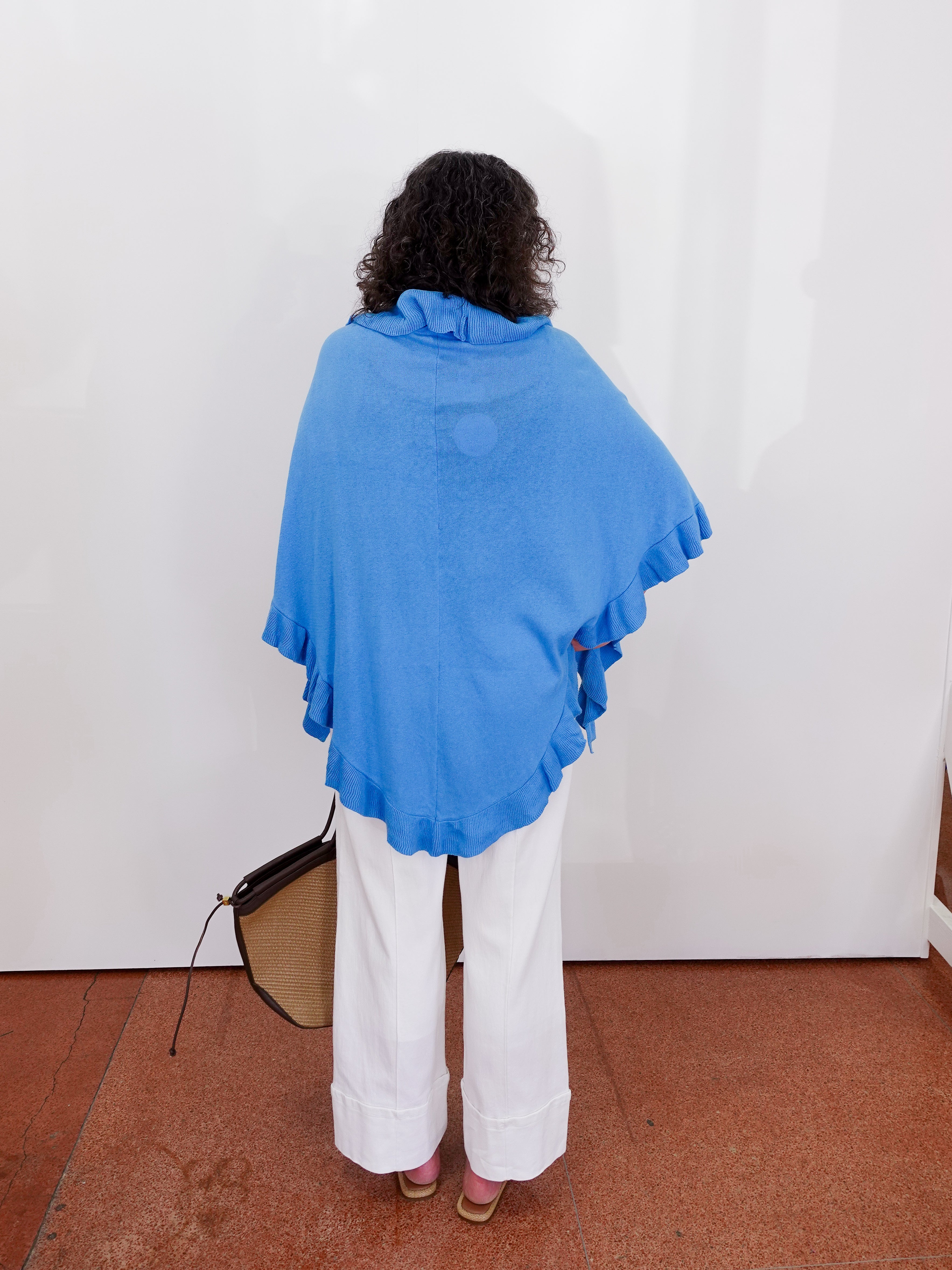 Person wearing a blue poncho and white pants against a white wall.