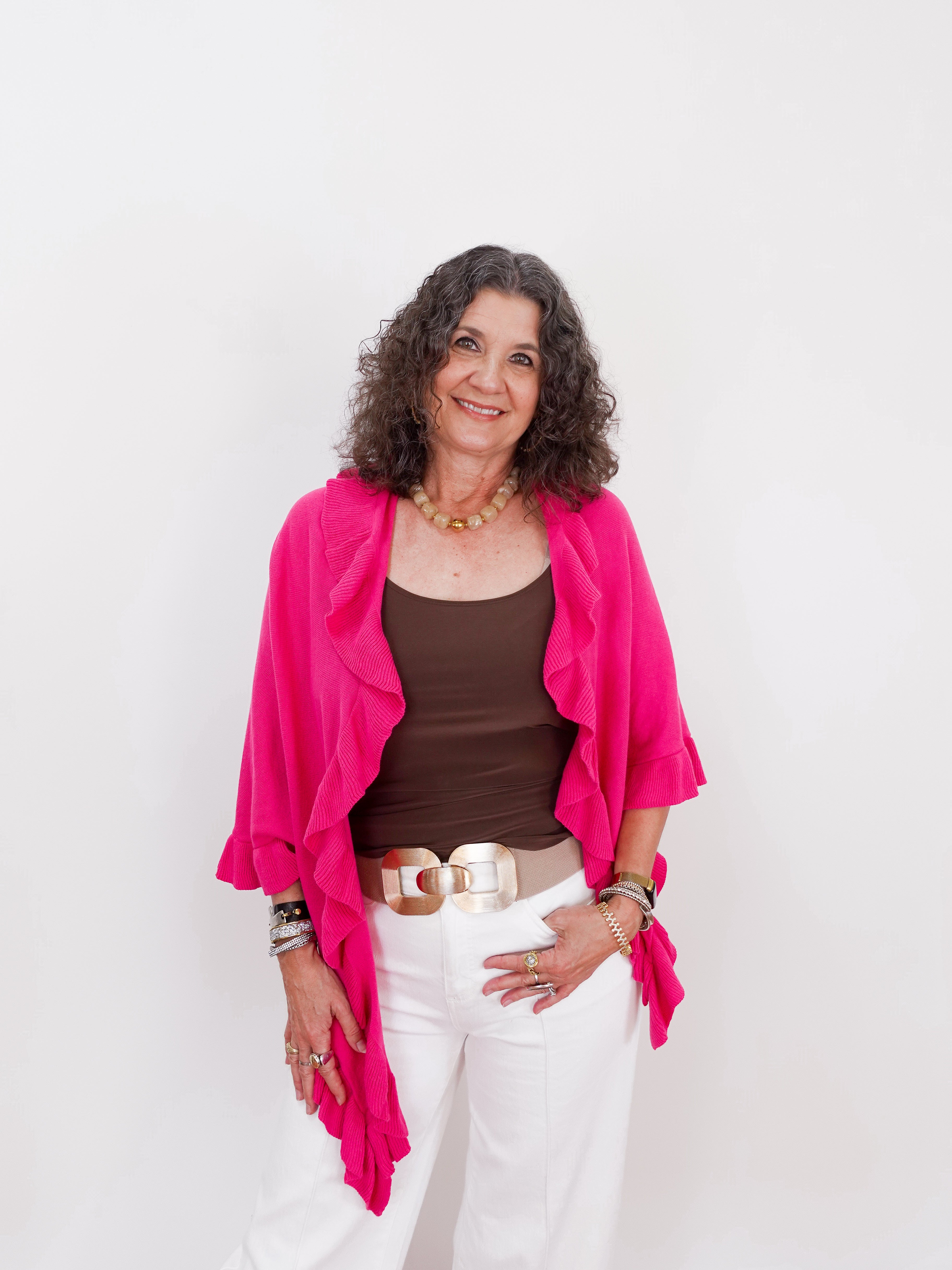 Woman wearing a bright pink cardigan over a brown top and white pants on a light gray background