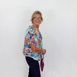 Woman wearing a colorful floral blouse and dark pants against a white wall.