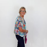 Woman wearing a colorful floral blouse and dark pants against a white wall.