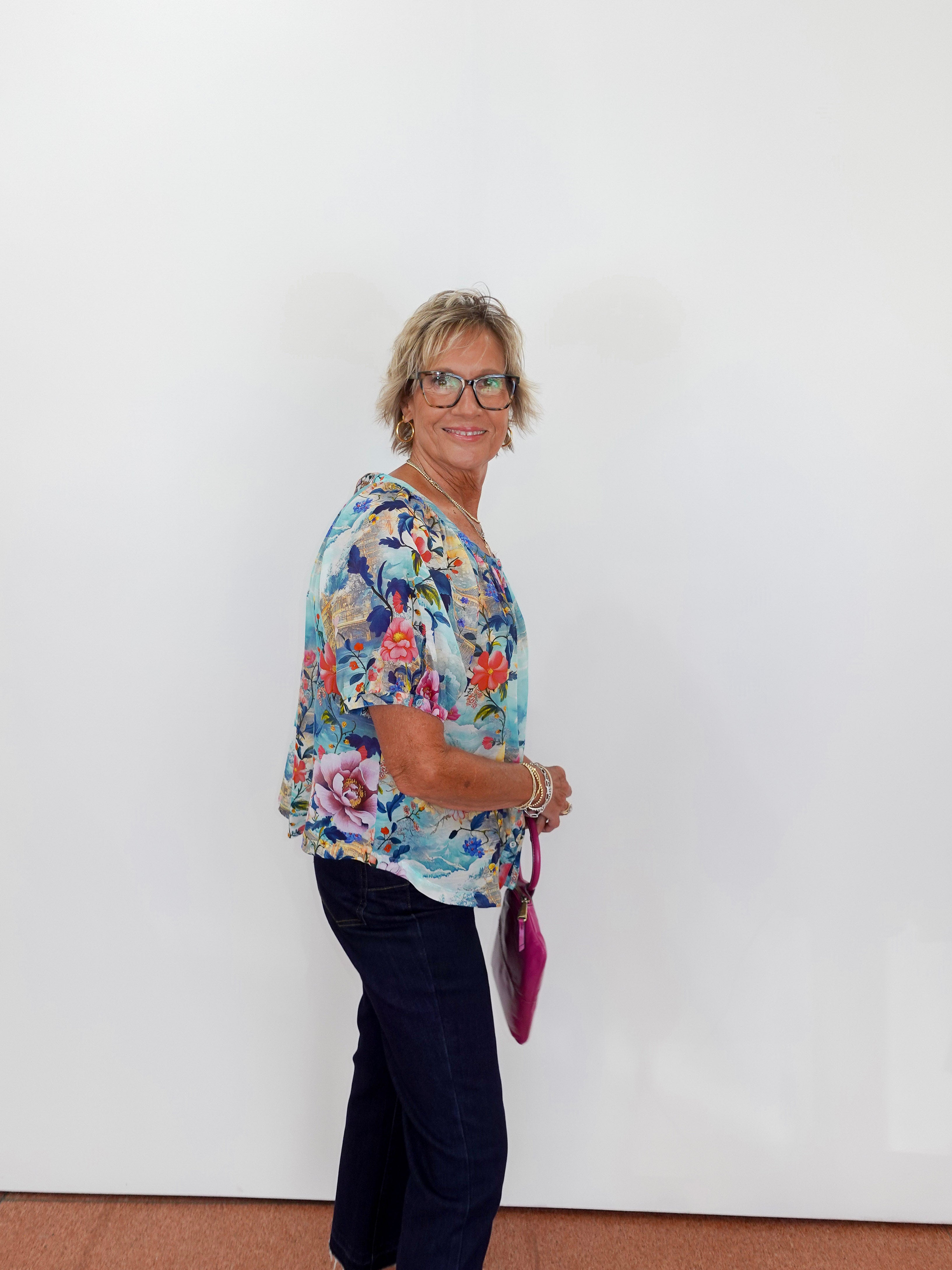 Woman wearing a colorful floral blouse and dark pants against a white wall.