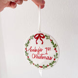 Decorative ornament with 'Baby's 1st Christmas' text and a red bow, held by a hand against a white background.