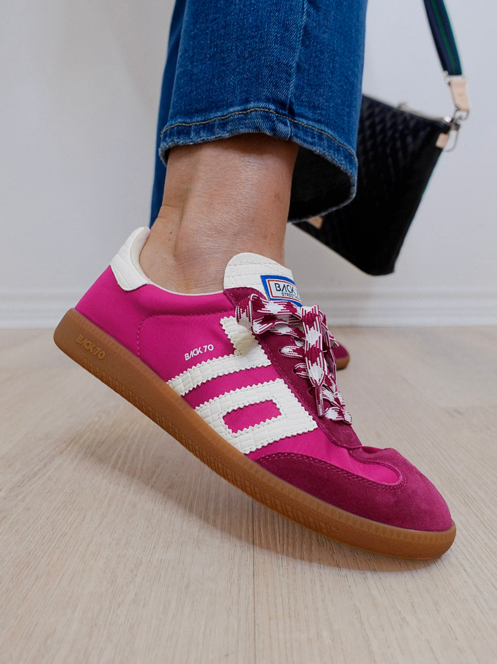 Pink Adidas sneaker with white stripes worn by a person on a light wooden floor.
