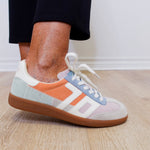 Multicolored sneaker with white laces worn indoors on a wooden floor.