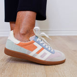 Multicolored sneaker with white laces worn indoors on a wooden floor.