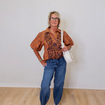 Woman wearing a patterned shirt, jeans, and white sneakers against a plain wall.