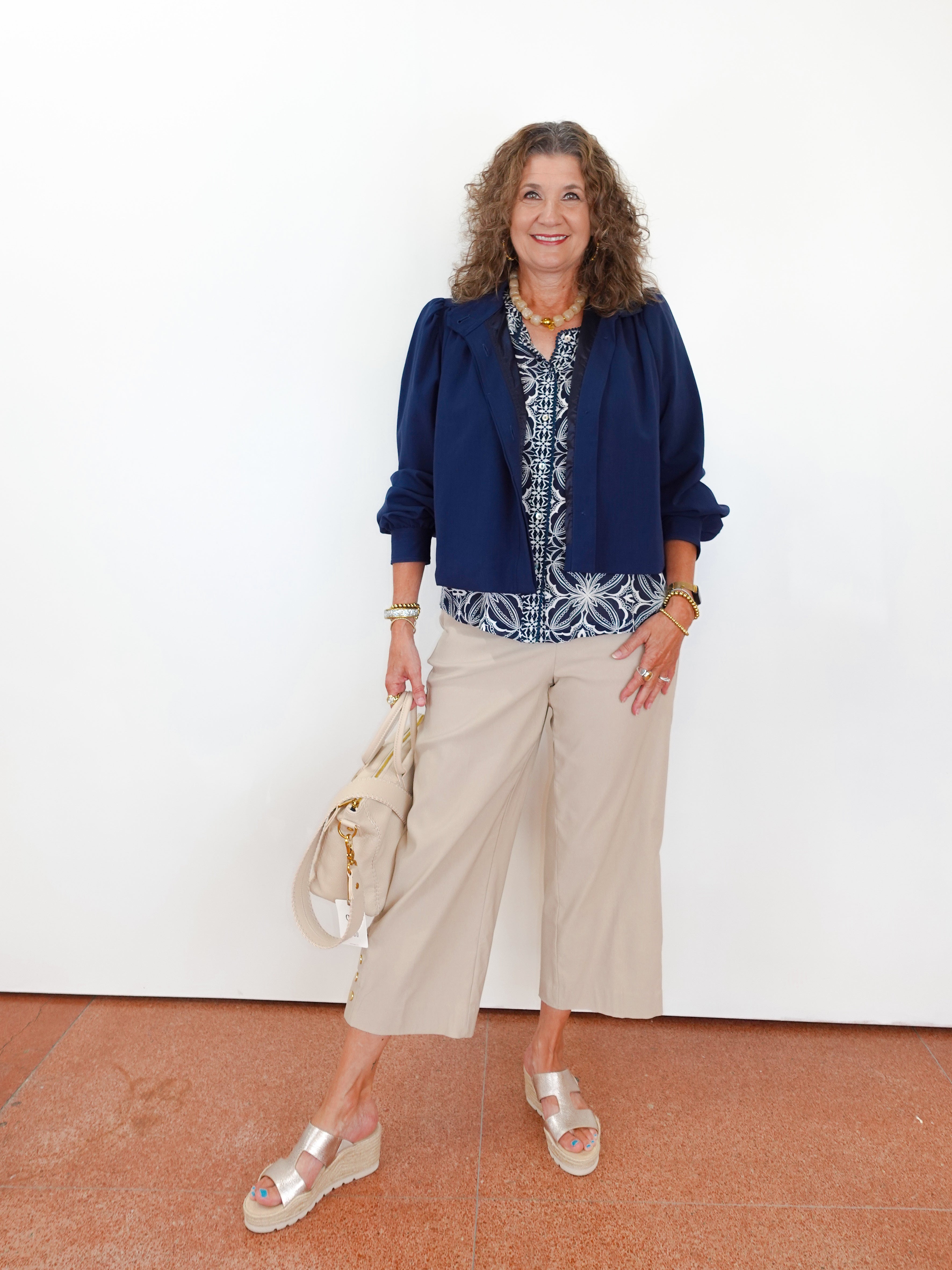 Woman wearing a blue jacket and beige pants against a white wall.