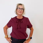 Woman wearing a burgundy blouse with a decorative bow and black pants against a white background