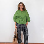 Woman wearing a green top and patterned pants standing against a white wall.