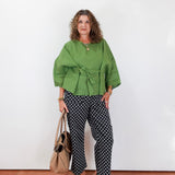 Woman wearing a green top and patterned pants standing against a white wall.