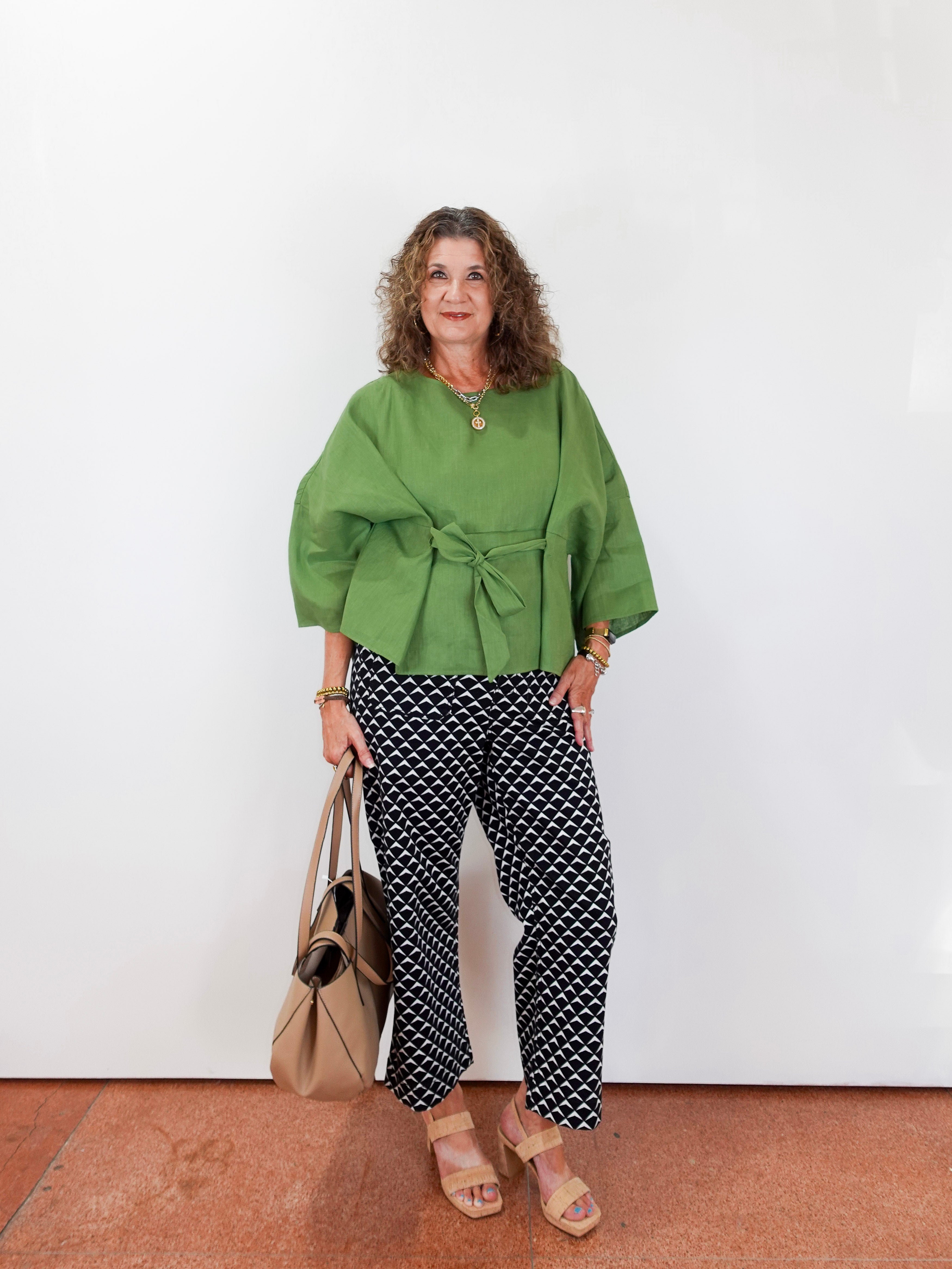 Woman wearing a green top and patterned pants standing against a white wall.