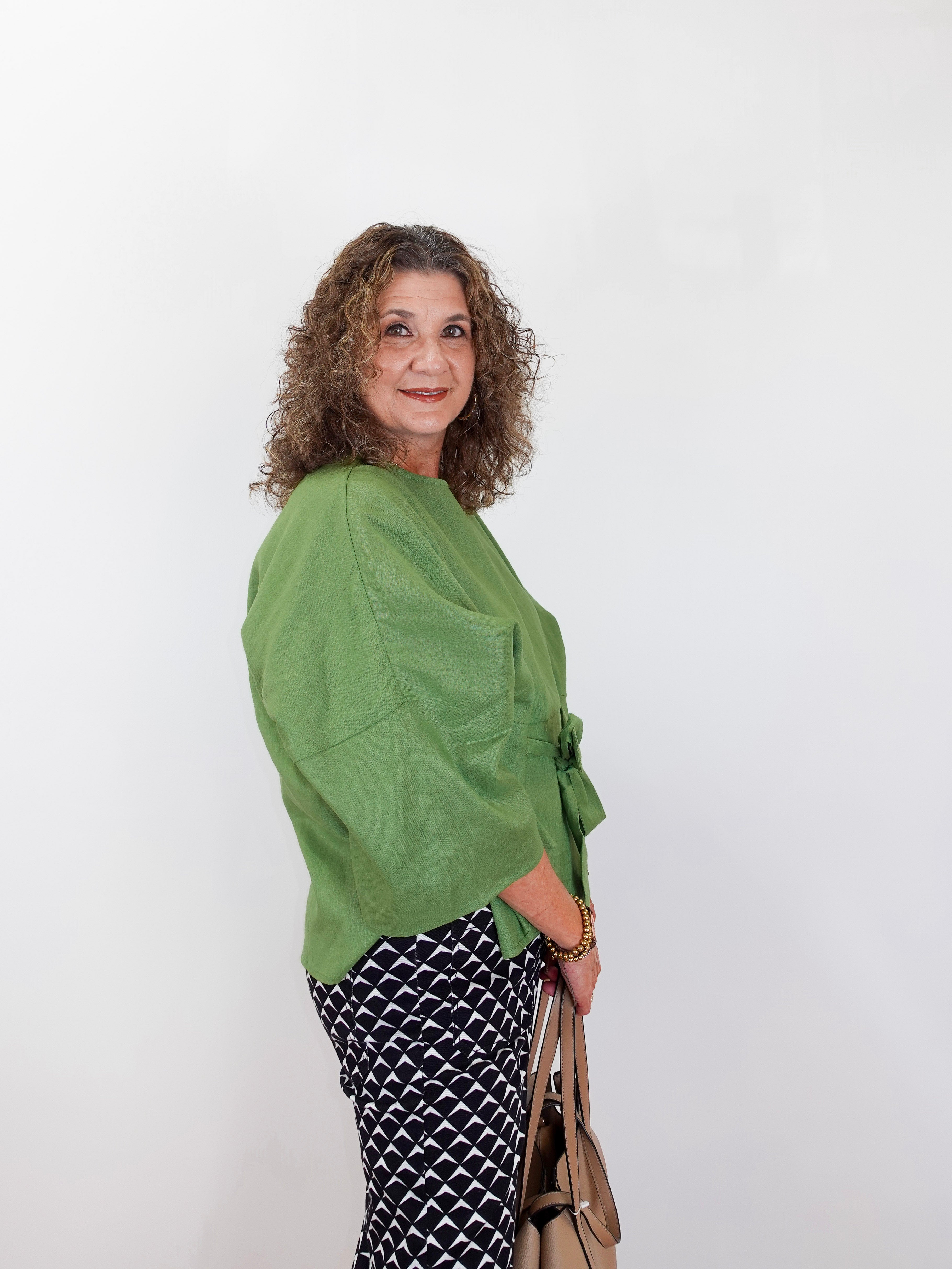 Woman wearing a green top and patterned pants holding a beige handbag against a white background