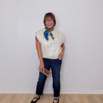 Person wearing a white blouse with a blue scarf, dark jeans, and black shoes against a white wall.
