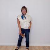 Person wearing a white blouse with a blue scarf, dark jeans, and black shoes against a white wall.