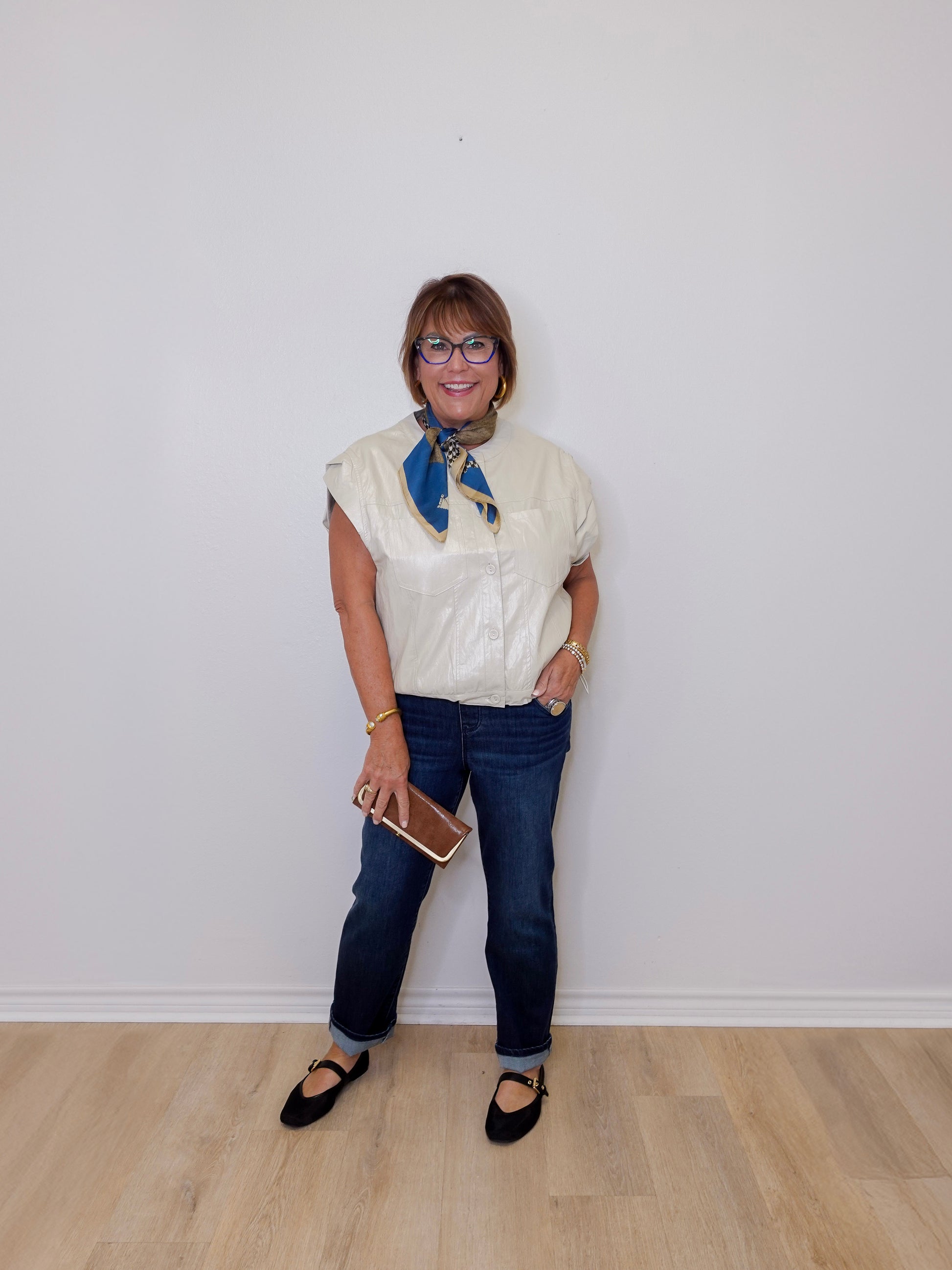 Person wearing a white blouse with a blue scarf, dark jeans, and black shoes against a white wall.
