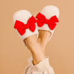 White slippers with red bows worn by a person on a beige background
