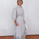 Woman wearing a light gray dress with floral patterns against a white wall.