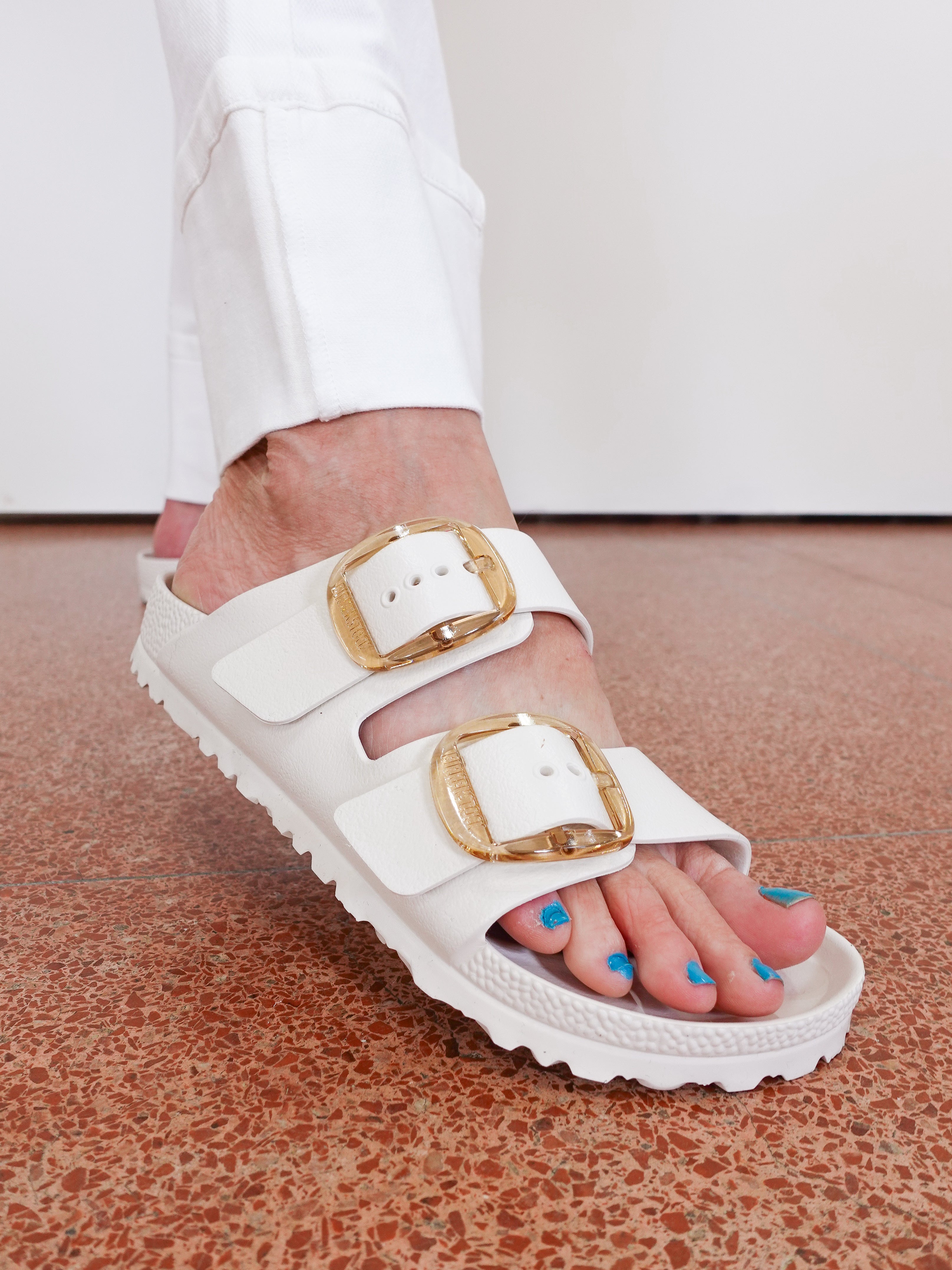 White sandals with gold buckles worn by a person on a terrazzo floor.