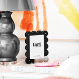Black scalloped photo frame with 'tart' branding on a shelf with books and a lamp.