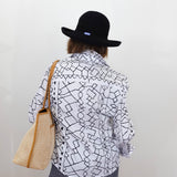 Person wearing a black and white patterned shirt, gray pants, and a black hat, holding a beige bag against a plain background.