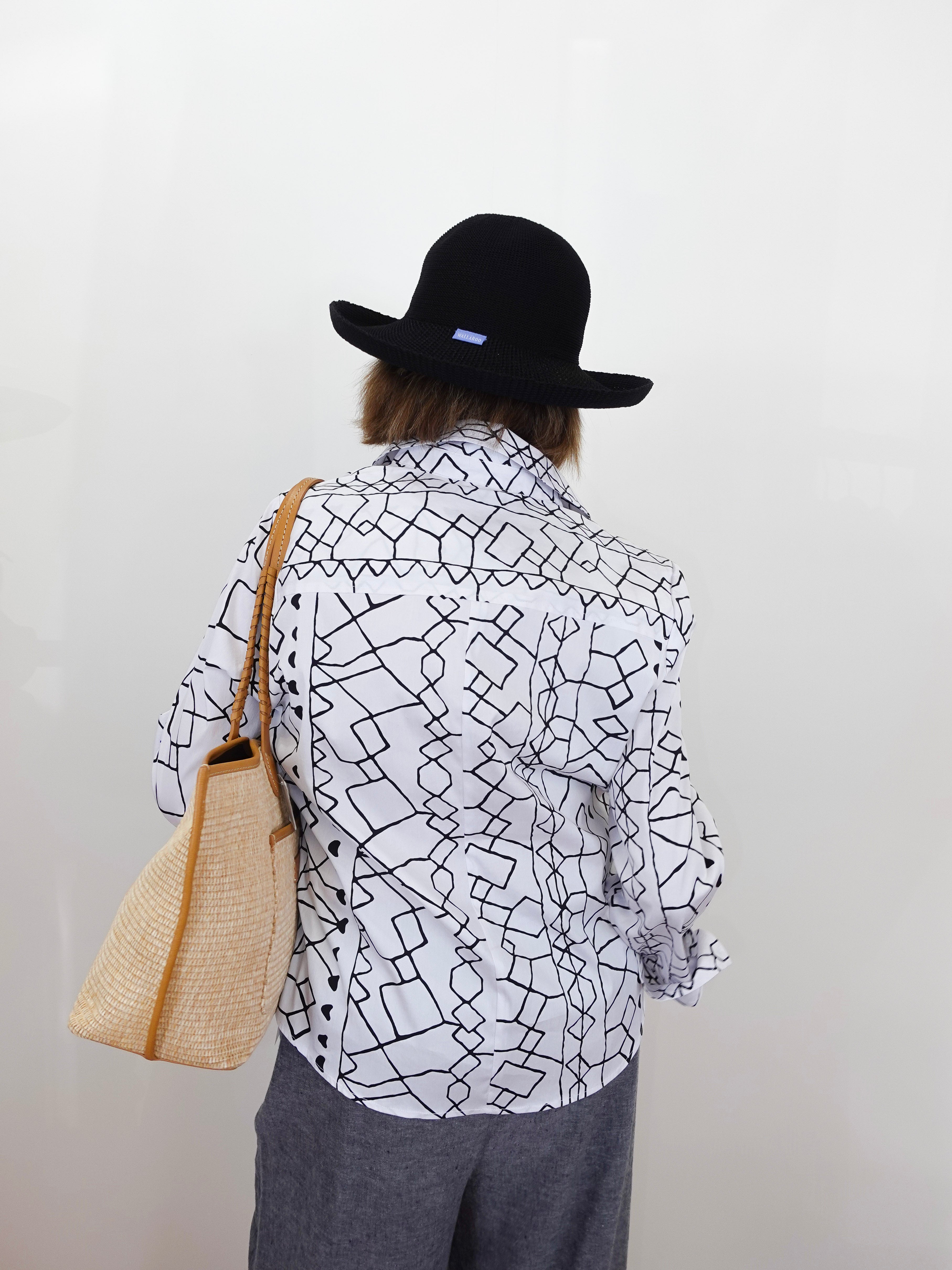 Person wearing a black and white patterned shirt, gray pants, and a black hat, holding a beige bag against a plain background.
