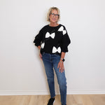 Person wearing a black sweater with white bow patterns and blue jeans on a plain background