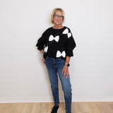 Person wearing a black sweater with white bow patterns and blue jeans on a plain background