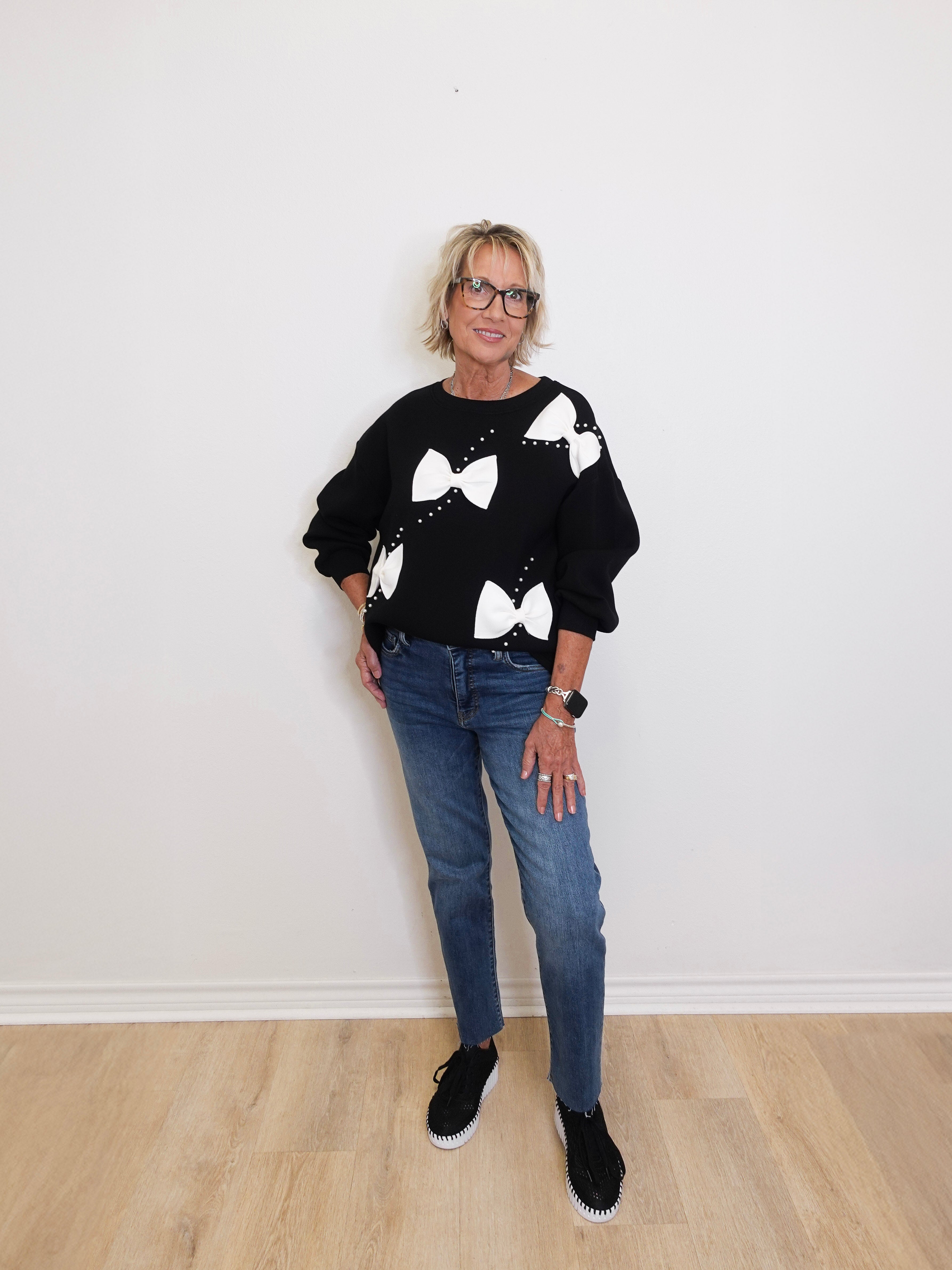 Person wearing a black sweater with white bow patterns and blue jeans on a plain background