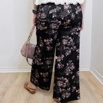 Person wearing black floral pants with a white lace top, standing indoors.