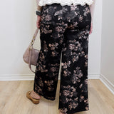 Person wearing black floral pants with a white lace top, standing indoors.