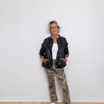 Person wearing a black leather jacket, white shirt, and camouflage pants against a white wall.