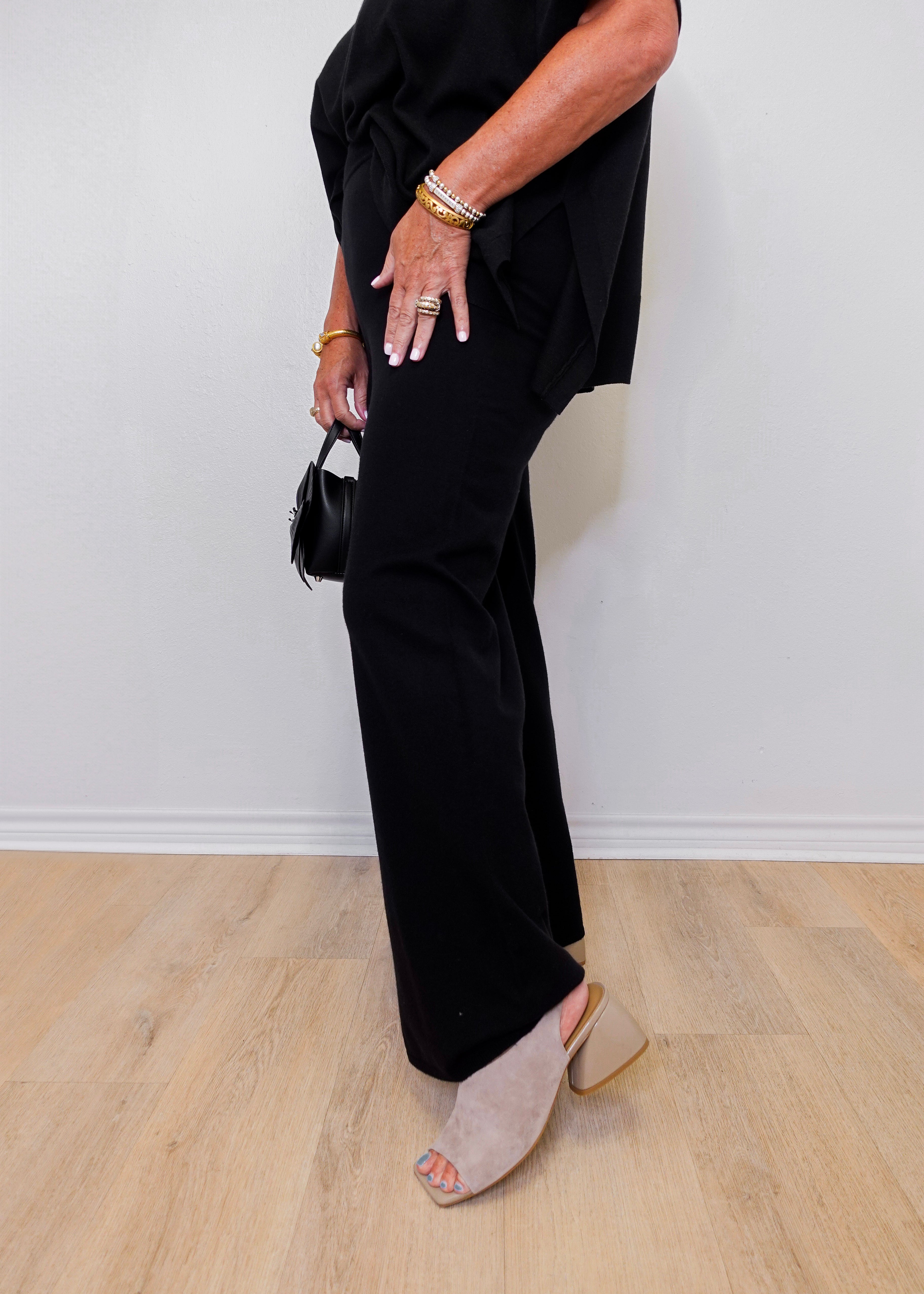 Person wearing a black outfit with wide-leg pants and open-toe shoes on a light wood floor.
