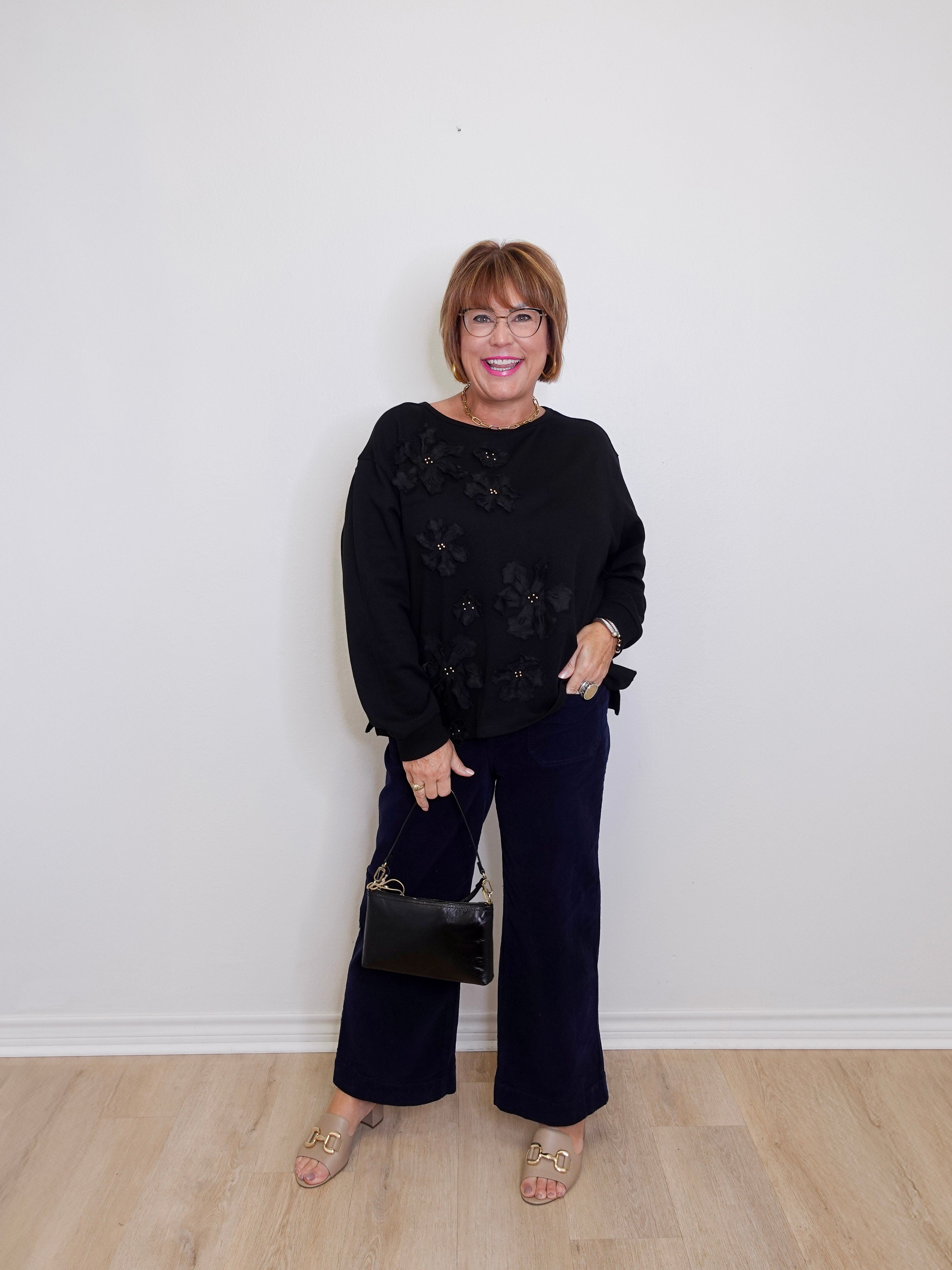 Woman wearing a black sweater and wide-leg pants against a white wall.