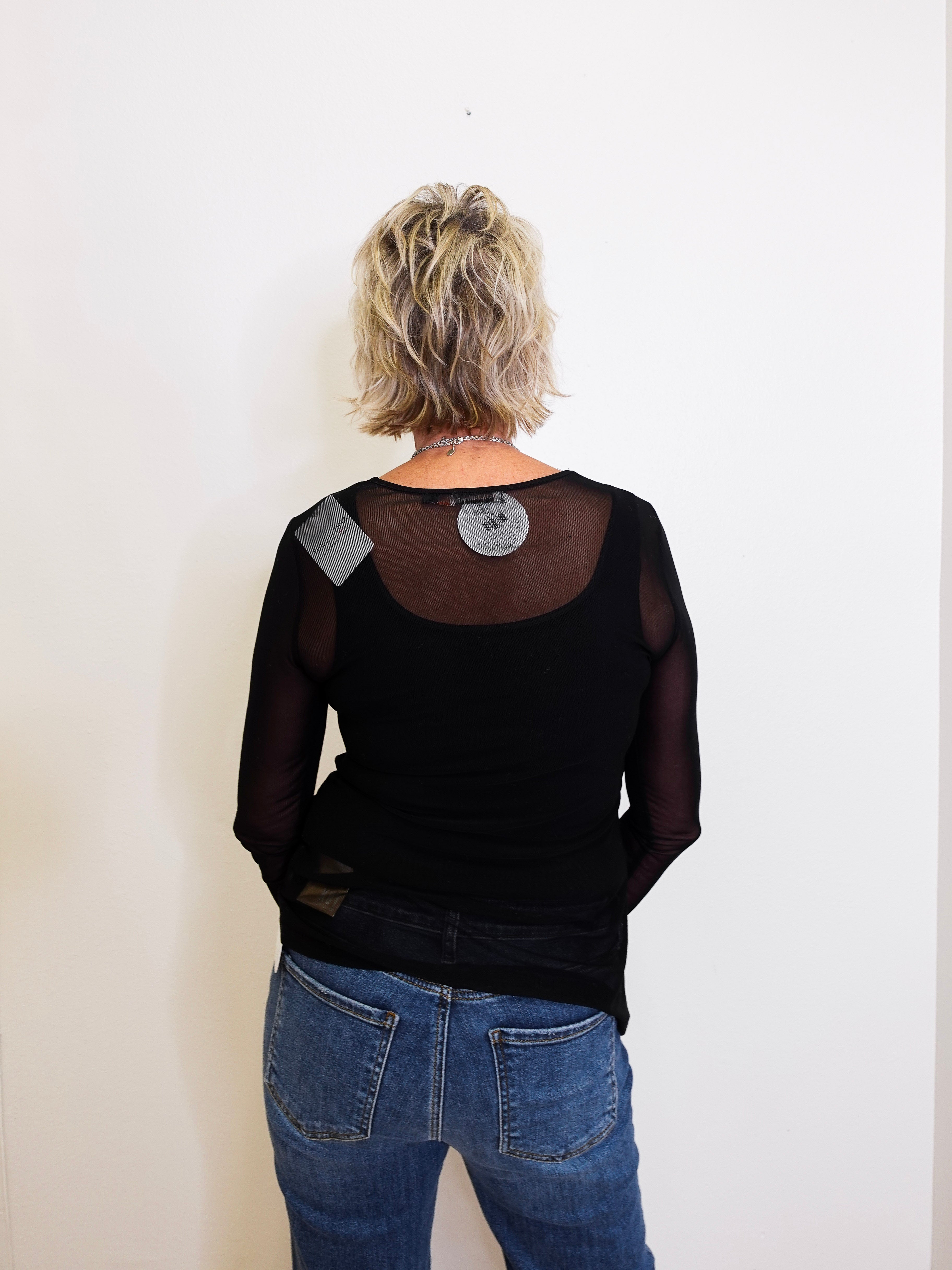 Person wearing a black long-sleeve top with sheer sleeves and blue jeans against a white background