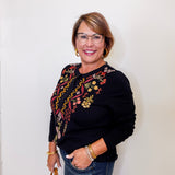 Woman wearing a black sweater with floral embroidery, holding a handbag against a white wall.