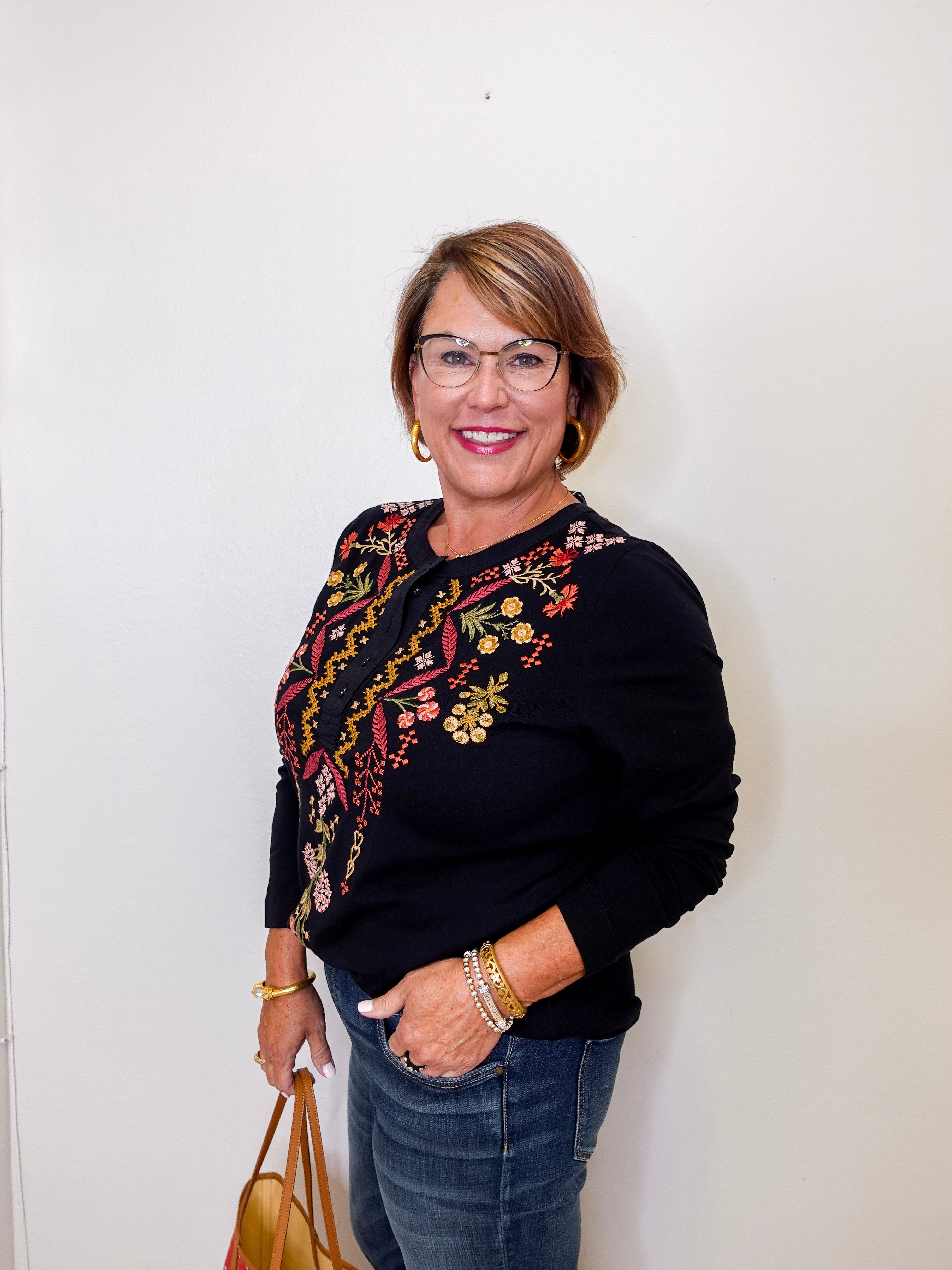 Woman wearing a black sweater with floral embroidery, holding a handbag against a white wall.