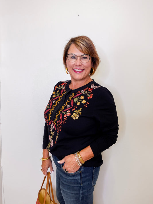 Woman wearing a black sweater with floral embroidery, holding a handbag against a white wall.