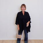 Woman wearing a black poncho and holding a patterned handbag against a white wall.