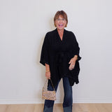 Woman wearing a black poncho and holding a patterned handbag against a white wall.