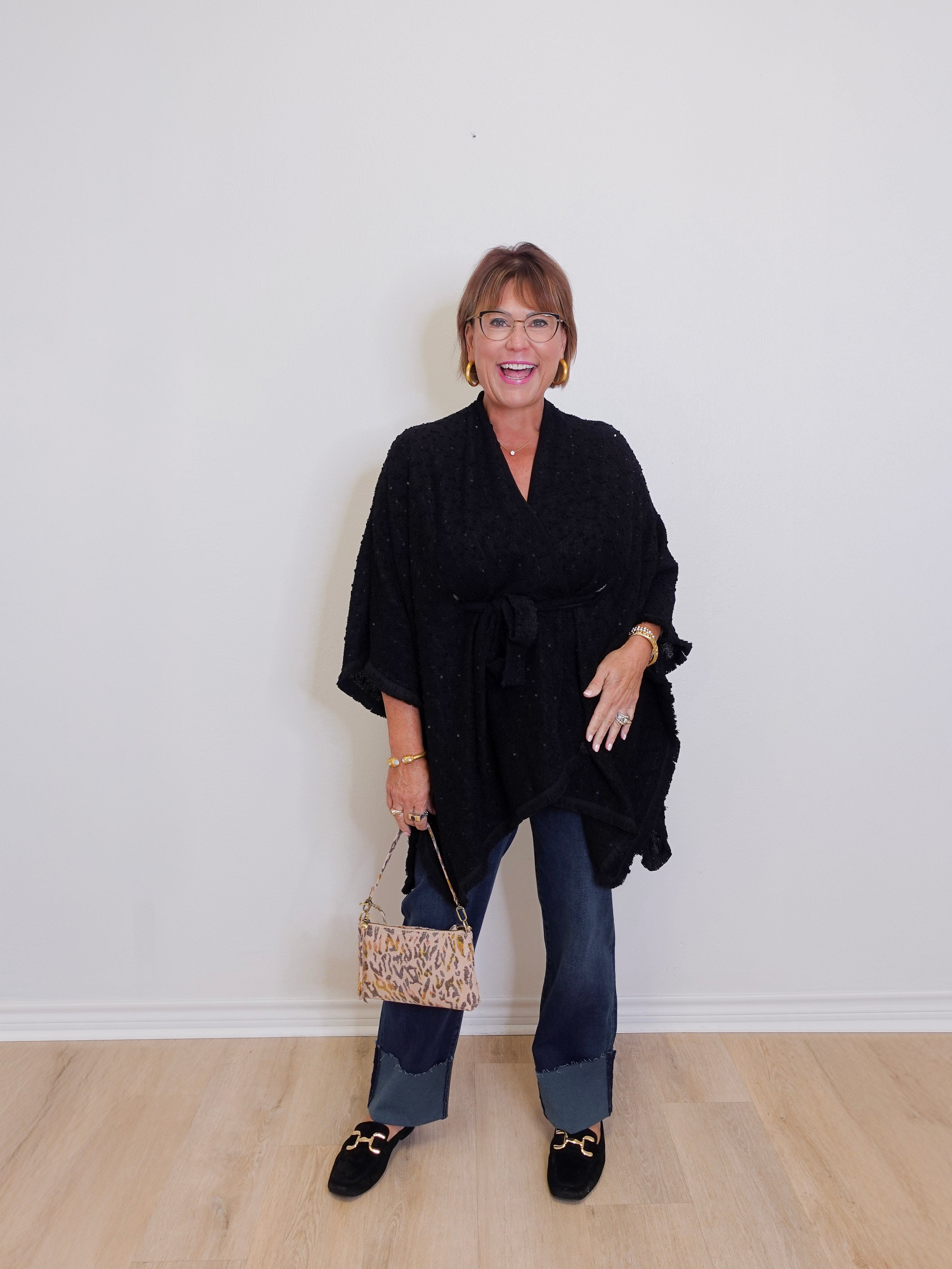 Woman wearing a black poncho and holding a patterned handbag against a white wall.