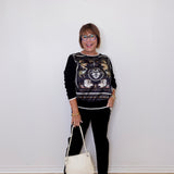Woman wearing a black patterned top and black pants, holding a white handbag against a white wall.