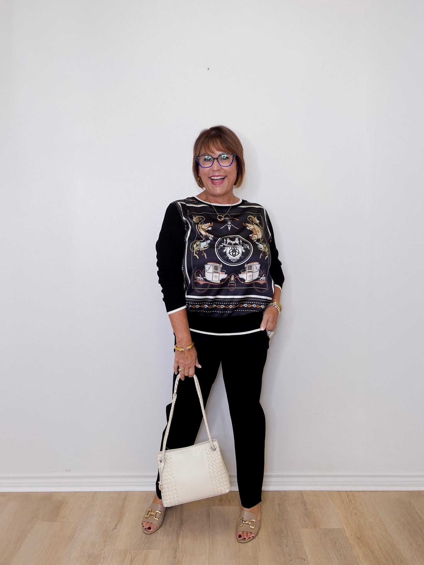 Woman wearing a black patterned top and black pants, holding a white handbag against a white wall.
