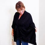 Woman wearing a black poncho and blue jeans against a white wall.