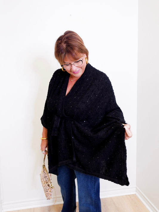Woman wearing a black poncho and blue jeans against a white wall.