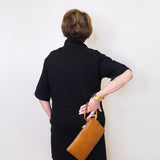 Person wearing a black dress holding a brown leather bag against a white wall.